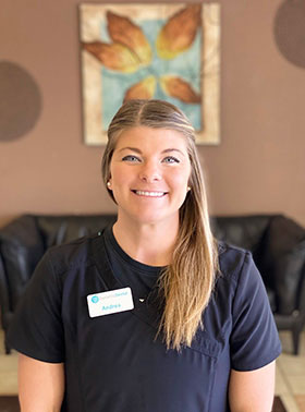 A smiling woman in a black and white uniform stands behind a desk, posing for a professional photo with a welcoming demeanor.