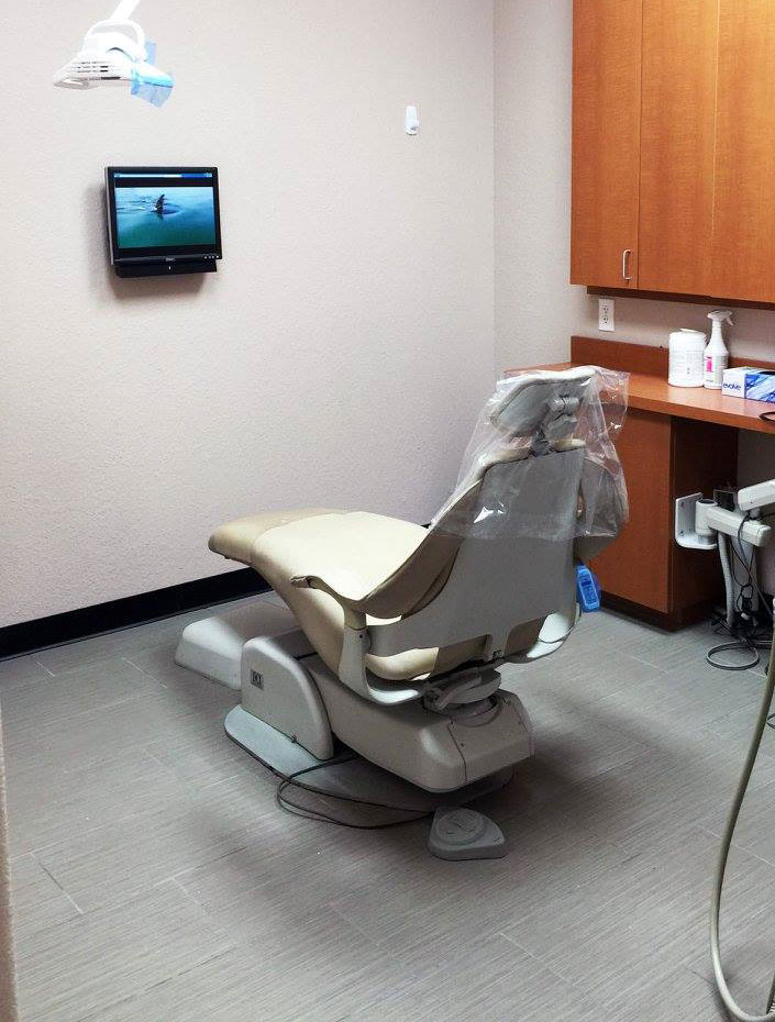 The image shows a dental office interior with a dental chair, overhead lighting, and a monitor displaying what appears to be an X-ray.