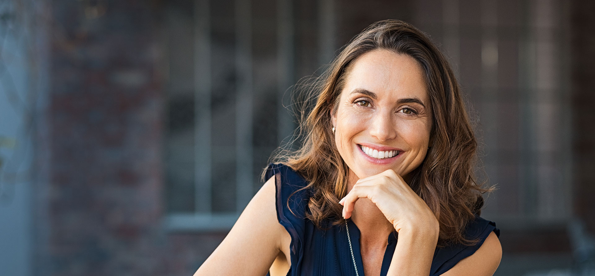 The image is a photograph of a woman with light skin, smiling at the camera. She appears to be in her late twenties or early thirties and has long hair. Her eyes are looking directly at the camera, and she is holding up her index finger near her mouth as if she s making a point or emphasizing something. The background is plain and light-colored, which suggests that this could be a stock photo used for various purposes such as advertising, personal branding, or lifestyle content.