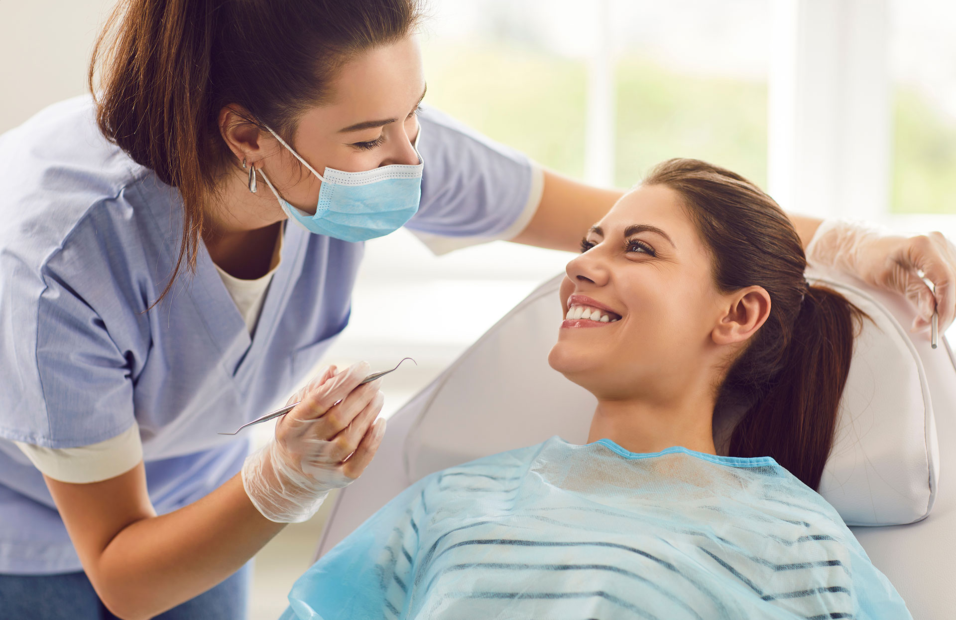 A dental hygienist is assisting a woman in a dental chair, with the dental hygienist smiling and holding a dental tool.