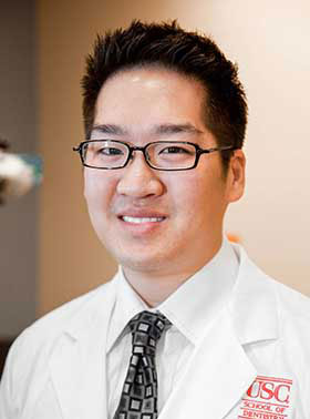 The image depicts a man wearing glasses, a white shirt, and a tie, standing in front of a microphone with a background that includes a logo.