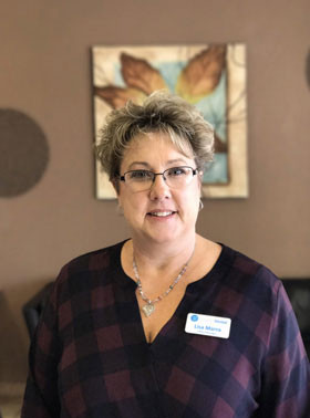 The image shows a woman standing in an indoor setting, possibly an office or lobby. She is wearing glasses and a dark top with a visible name tag.