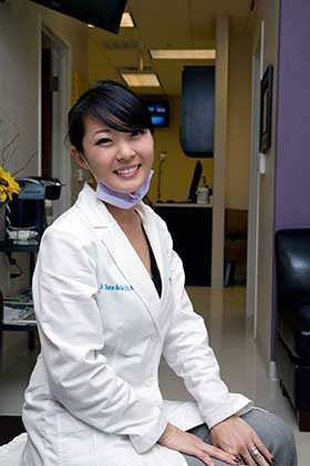 A woman wearing a white lab coat and mask is smiling at the camera, seated in a dental office setting.