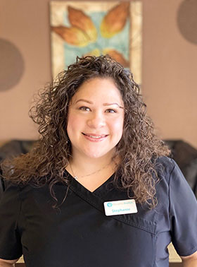 A woman in a black shirt and name tag, standing in front of a wall with a decorative element, smiling at the camera.