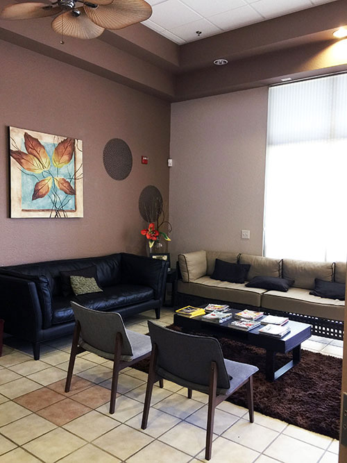 The image shows an interior view of a waiting room with a modern design, featuring a couch, chairs, a coffee table, and decorative elements such as framed artwork and a potted plant.
