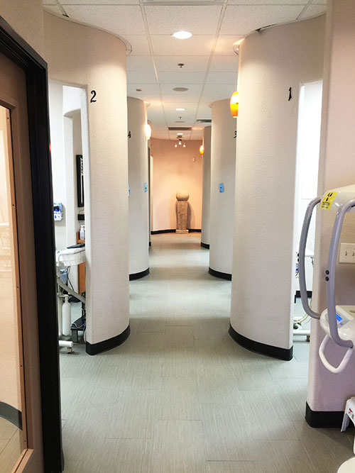 The image shows an interior hallway in a medical facility, with white walls and flooring, leading to a room with visible medical equipment.