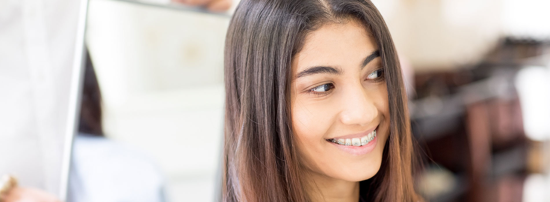 A woman smiling at the camera, with her reflection visible in a mirror behind her.