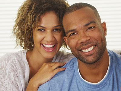 The image depicts a man and woman smiling at the camera, with the man wearing a short beard and the woman with curly hair. They appear to be in a happy and affectionate pose, suggesting a close relationship between them.