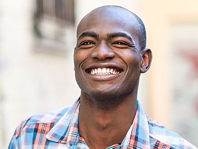 A young man with a broad smile, wearing a blue and white plaid shirt, against an outdoor background.