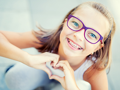 A young girl with glasses and a heart-shaped hand gesture is smiling at the camera.